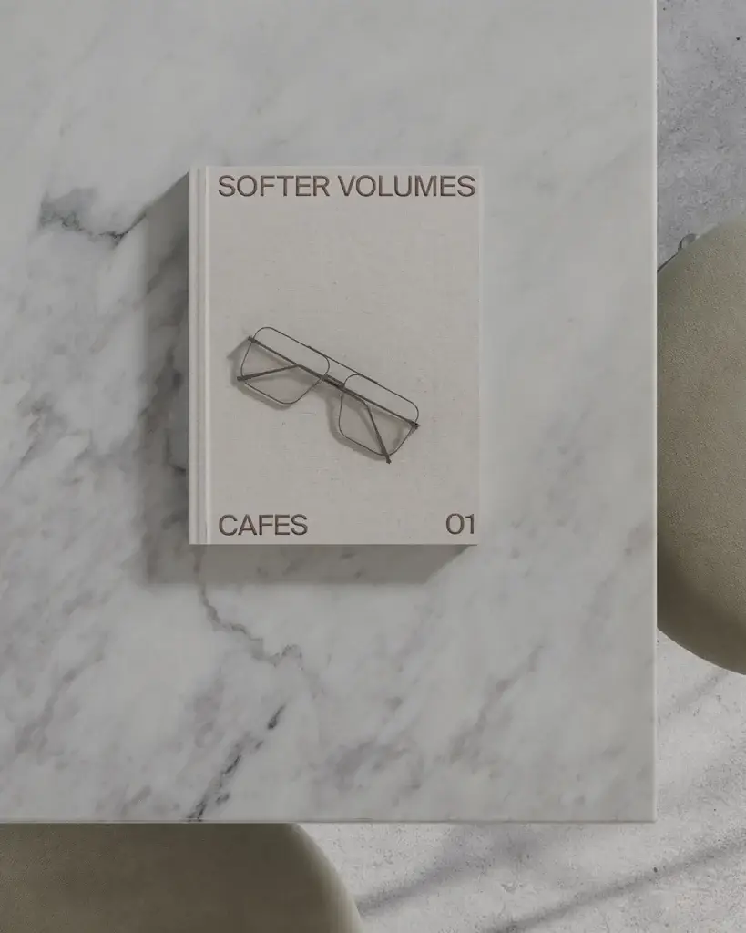Beige hardcover book titled Softer Volumes Cafes 01 with wire-frame glasses on its cover, placed on a marble surface.