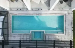 Aerial view of a modern rectangular pool with turquoise water and a pale gray surrounding deck, with glass railing and seating along the edge.