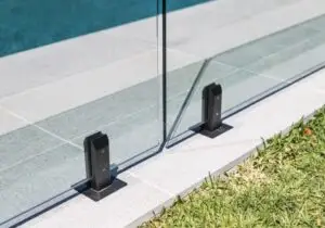 Glass pool railing supported by black metal standoffs anchored to a concrete deck along a pool edge, with grass nearby.