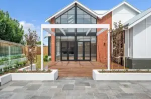 Modern brick house with large glass doors and a wooden deck at the entrance, with planters and young trees on both sides.