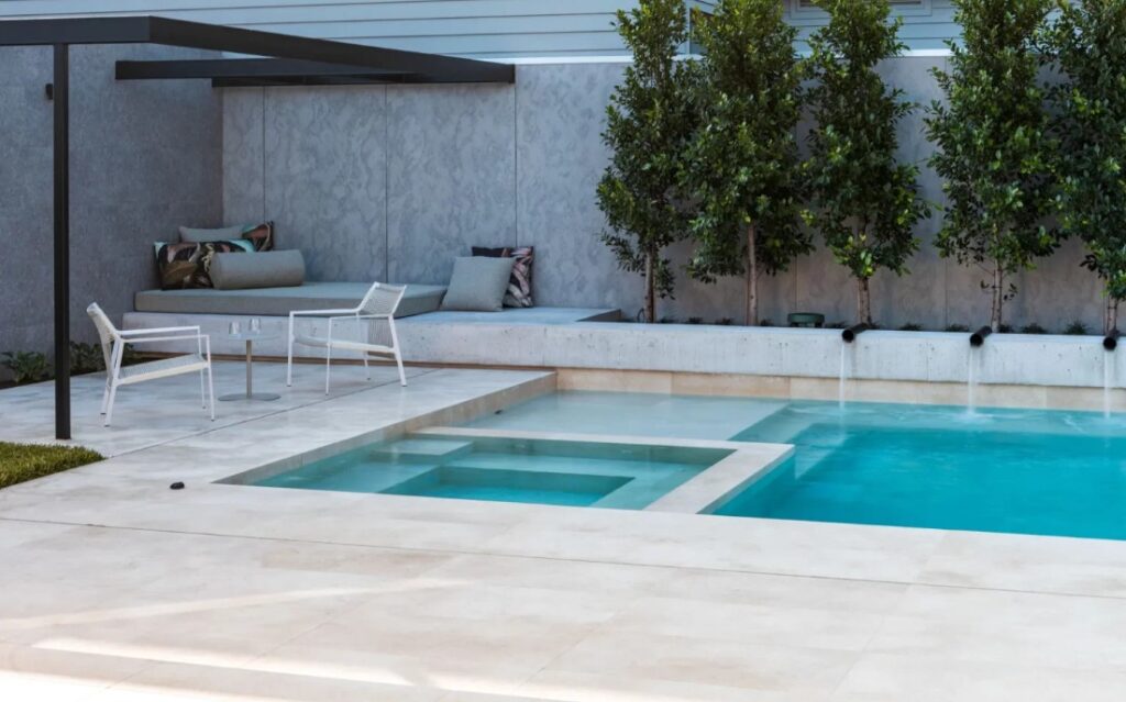 Poolside lounge area with white chairs and a glass table under a shaded canopy next to a gray wall and cushions.