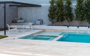 Poolside lounge area with white chairs and a glass table under a shaded canopy next to a gray wall and cushions.