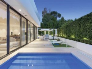 Modern glass-walled house with a rectangular blue pool in the foreground and a spacious white tiled patio beyond.