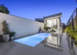 Modern backyard with a rectangular illuminated pool, white deck, and glass-railed lounge area beside a contemporary home during dusk.