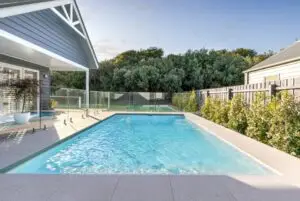 Rectangular in-ground pool in a backyard with a glass fence, beige deck tiles, and a blue house with a covered patio outside.