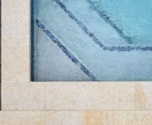 Top-down view of a swimming pool corner with beige stone coping and a dark blue tile edge, a diagonal line guiding toward the water.