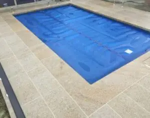 Outdoor rectangular pool covered with a blue safety cover, surrounded by beige tile decking.