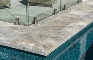 Close-up of a pool edge with gray stone coping and a glass railing, blue tiled pool wall below.