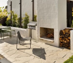Outdoor patio with a built-in brick barbecue, woodpile, and a single gray chair on irregular stone paving.