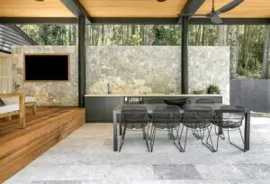 Outdoor covered patio with a stone wall, wood decking, and a long dining table surrounded by eight black wire chairs under a wooden ceiling. Nearby is a modern outdoor kitchen counter.