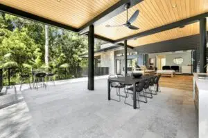 Covered outdoor dining area with a long black table and eight chairs, opening to a tropical garden and interior living space beyond.
