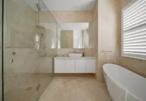 Modern beige-tiled bathroom with a glass-walled shower, white vanity with a vessel sink, a freestanding tub, and a window with blinds.