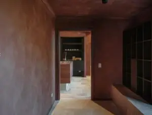 Brown-toned hallway opening to a workspace with a wooden counter and computer monitor. Dark shelves line the back wall; tiled floor visible.