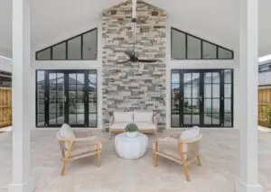 Covered outdoor patio with beige wicker chairs and a small sofa arranged around a white round table, centered by a stacked-stone wall and a ceiling fan; black-framed glass doors on either side.
