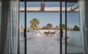 Sliding glass doors open to a sunny rooftop terrace with a round table and chairs and a palm tree in the background.