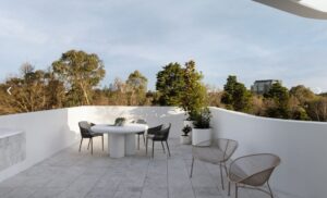 Modern white rooftop terrace with round white table and black chairs, potted plants, and a city skyline in the distance.
