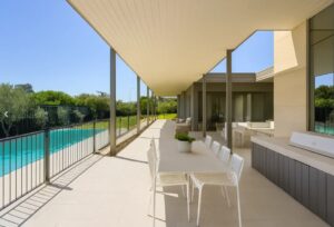 Modern outdoor patio with a long white dining table and chairs under a covered walkway beside a pool on the left.