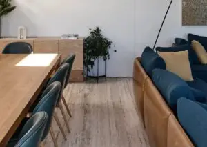 Wooden dining table with teal chairs in a bright open living area, plant and sideboard in the background, blue sofa along the right wall.