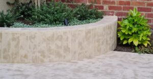 Curved beige-tiled planter with green shrubs and a small spotlight, set against a brick wall and tiled patio.
