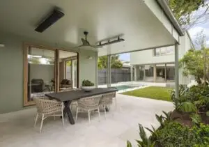 Outdoor covered patio with a dark rectangular dining table and six woven chairs, overlooking a modern backyard with a pool and lawn.