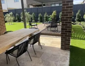 Outdoor backyard patio with a long wooden dining table and black woven chairs on a tiled floor, surrounded by a green lawn and shrubbery fence in the backyard.